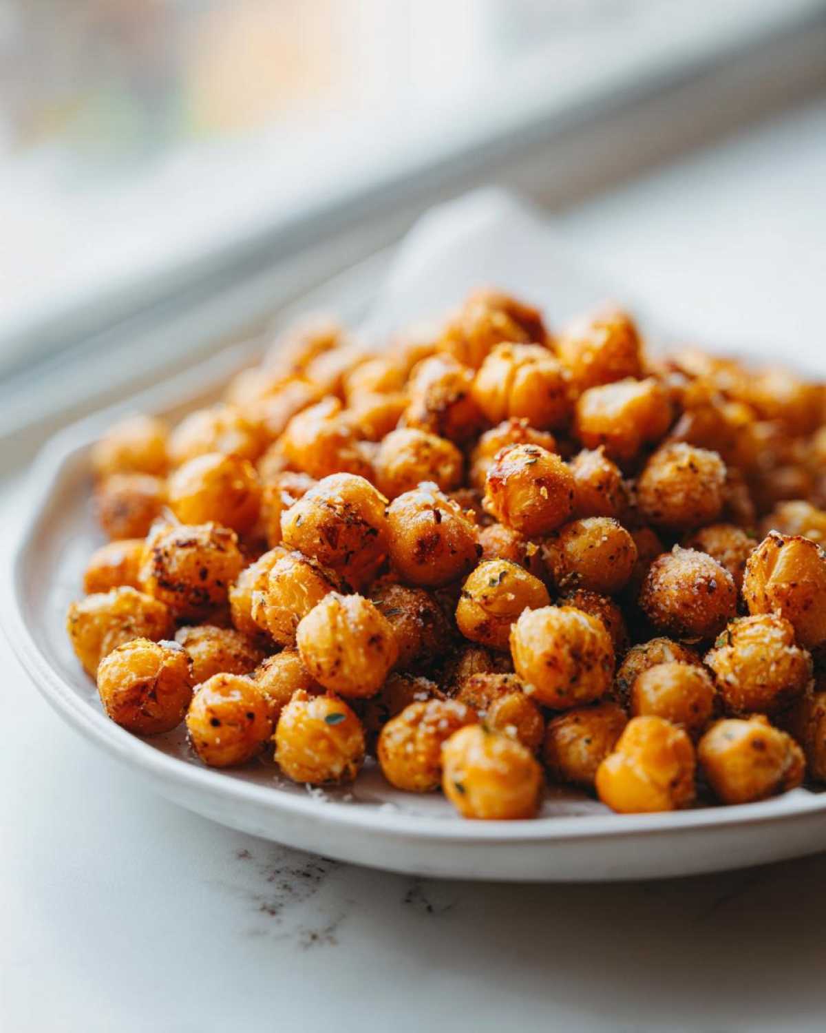 A close-up of crispy, seasoned Air Fryer Ranch Chickpeas piled high on a white plate near a window.