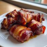 A close-up of several glossy, caramelized Apple Butter Bacon Wrapped Smokies piled on a white plate.