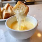 A piece of toasted bread is dipped into a white bowl of Authentic White Wine Gruyère Fondue, showing long cheese pulls.