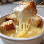 A piece of toasted bread is lifted from a white bowl filled with creamy Authentic White Wine Gruyère Fondue, showing long cheese strings.
