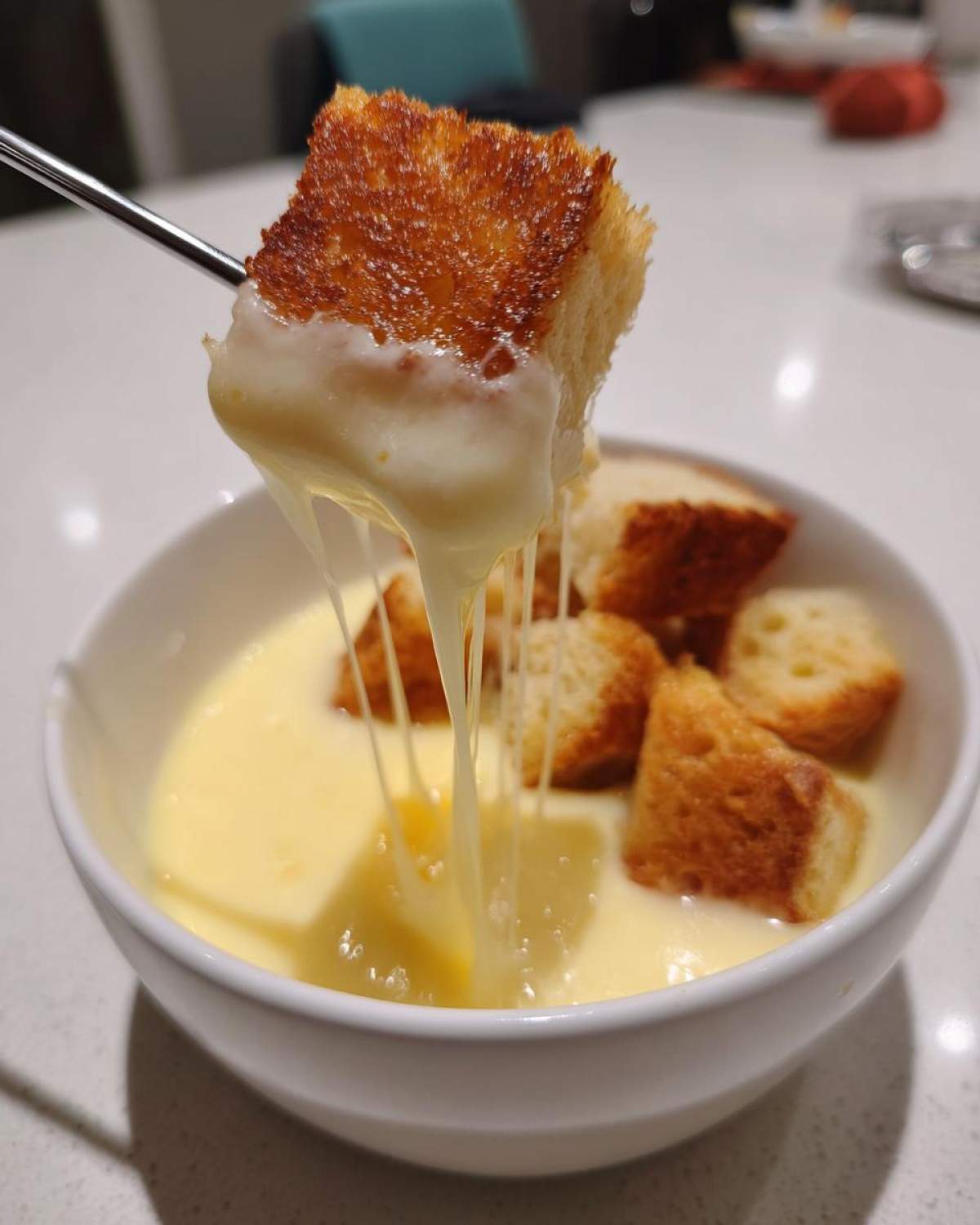 A piece of toasted bread is lifted from a bowl of creamy Authentic White Wine Gruyère Fondue, showing long, stretchy cheese pulls.