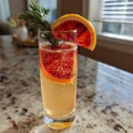 A tall glass of Blood Orange Mimosa with Rosemary, garnished with a blood orange wheel and fresh rosemary sprig.