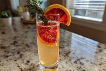 A tall glass of Blood Orange Mimosa with Rosemary, garnished with a blood orange wheel and fresh rosemary sprig.