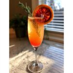 A vibrant Blood Orange Mimosa with Rosemary garnished with a blood orange slice and rosemary sprig.