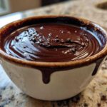 Close-up of rich, glossy Bourbon Dark Chocolate Fondue topped with chocolate shavings in a rustic white and brown bowl.