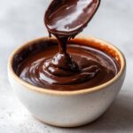 Close-up of thick, glossy Bourbon Dark Chocolate Fondue being poured from a spoon into a speckled white bowl.