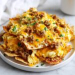 A plate piled high with Breakfast Nachos with Sausage and Scrambled Eggs, topped with melted cheddar cheese and parsley.