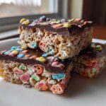 Three stacked Chocolate Dipped Lucky Charms Marshmallow Bars topped with colorful cereal pieces on a white plate.