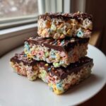 Three stacked Chocolate Dipped Lucky Charms Marshmallow Bars topped with colorful sprinkles on a white plate.
