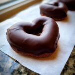 Close-up of a heart-shaped treat, likely a donut or pastry, generously coated in dark chocolate, resembling Chocolate Dipped Pear Hearts.