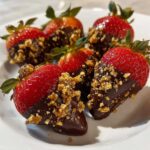 Five fresh strawberries dipped in dark chocolate and generously coated with crunchy toffee pieces, ready to eat.