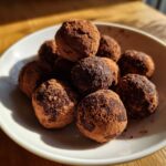 A close-up of several rich Date and Almond Chocolate Truffles piled in a white bowl, heavily dusted with cocoa powder.