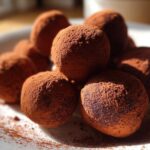 Close-up of several rich Date and Almond Chocolate Truffles heavily dusted with cocoa powder on a white plate.