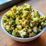 Close-up of fluffy popcorn generously coated with green Dill Pickle Seasoned Popcorn powder in a white bowl.