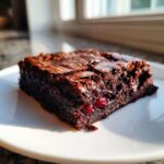 A close-up of a fudgy Dark Chocolate Cherry Brownies slice on a white plate, showing visible cherries.