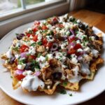A plate piled high with Greek Chicken Nachos featuring shredded chicken, feta, tomatoes, olives, and tzatziki sauce on pita chips.