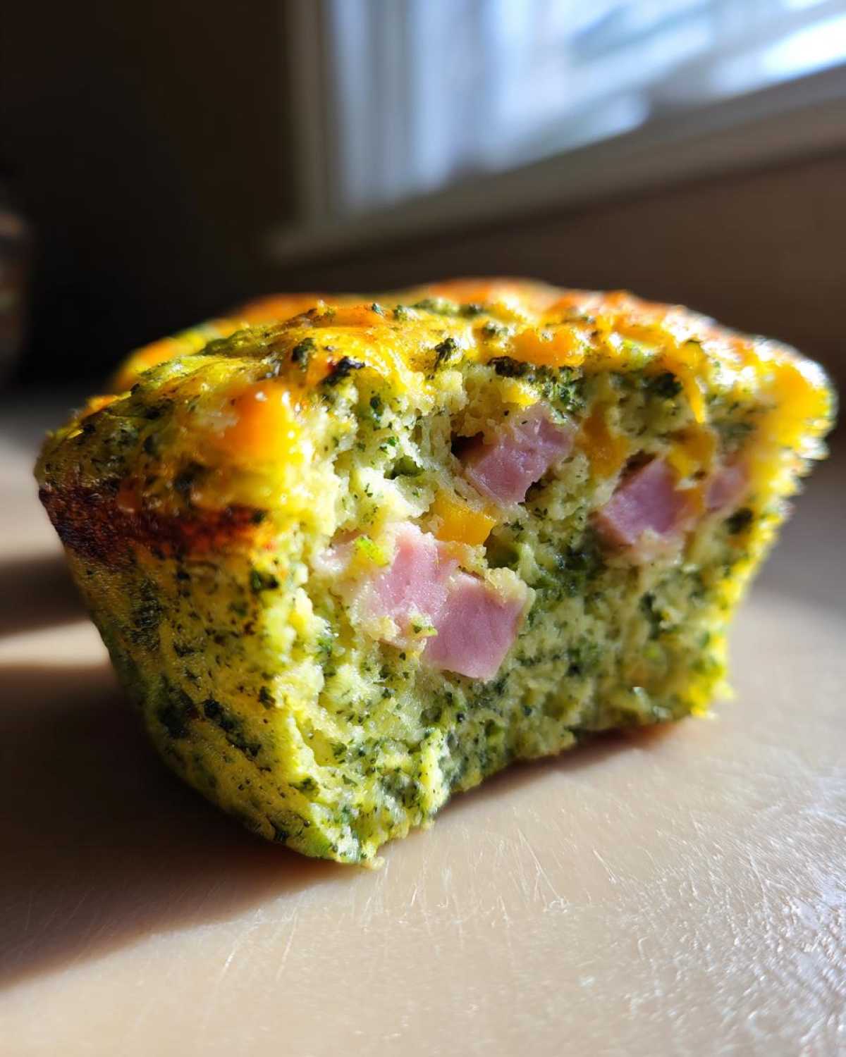 Close-up of a Green Eggs and Ham Savory Muffin cut in half, showing green flecks, chunks of pink ham, and melted cheese.