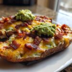 Two halves of baked potato skins loaded with melted cheese, bacon bits, and topped with guacamole, ready to eat.