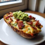 A single, loaded potato skin topped with melted cheese, crispy bacon bits, and a dollop of guacamole, ready to eat.