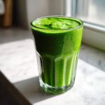 A tall glass filled with a thick, vibrant Healthy Green Spinach Milkshake sitting on a white marble surface near a window.