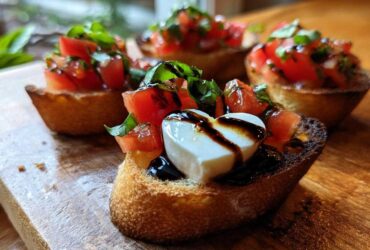 Close-up of Heart-Shaped Tomato & Mozzarella Bruschetta topped with balsamic glaze and basil on a wooden board.
