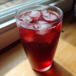 A tall glass filled with vibrant red Hibiscus Iced Tea with Honey and ice cubes, showing condensation.