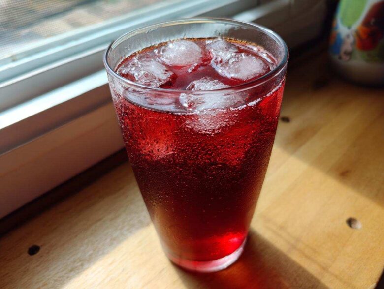 A tall glass filled with vibrant red Hibiscus Iced Tea with Honey and ice cubes, showing condensation.