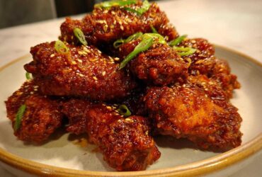 A pile of glossy, dark red Korean Fried Chicken Wings coated in sauce, sprinkled with sesame seeds and green onions.