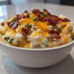 A close-up of a white bowl filled with creamy Loaded Baked Potato Dip, topped with shredded cheddar, bacon bits, and green onions.