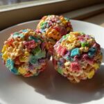 Three brightly colored Lucky Charms Popcorn Balls drizzled with white icing, sitting on a white plate near a window.