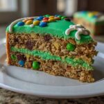A slice of Monster Cookie Football Cake showing layers of cookie base, green frosting, chocolate chips, and M&Ms on a white plate.