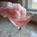 A pink Cotton Candy Martini in a coupe glass, topped with a large puff of pink cotton candy.
