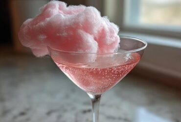 A pink Cotton Candy Martini in a coupe glass, topped with a large puff of pink cotton candy.