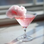 A sparkling pink cocktail in a martini glass, garnished with a puff of pink cotton candy, representing a Pink Cotton Candy Martini.