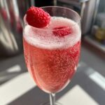 Close-up of a bubbly, pink French 75 Cocktail garnished with a fresh raspberry on the rim.
