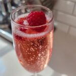 Close-up of a fizzy Pink French 75 Cocktail garnished with a fresh raspberry in a flute glass.