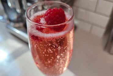 Close-up of a fizzy Pink French 75 Cocktail garnished with a fresh raspberry in a flute glass.