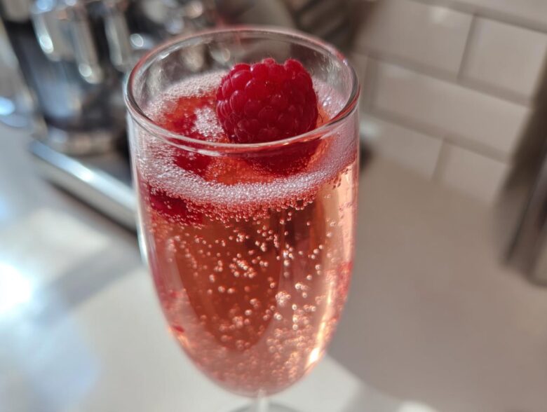 Close-up of a fizzy Pink French 75 Cocktail garnished with a fresh raspberry in a flute glass.