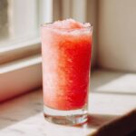 A tall glass filled with a bright red, icy Pink Lemonade Fruit Slushie sitting on a sunny marble surface.