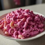 A generous serving of vibrant Pink Macaroni and Cheese, coated in a bright pink sauce, sits on a white plate.