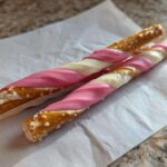 Two Pink White Chocolate Dipped Pretzel Rods with visible salt crystals resting on white parchment paper.