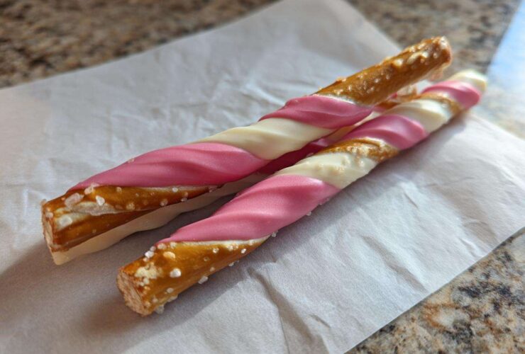 Two Pink White Chocolate Dipped Pretzel Rods with visible salt crystals resting on white parchment paper.