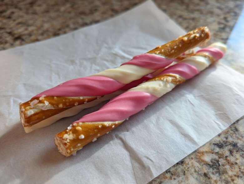 Two Pink White Chocolate Dipped Pretzel Rods with visible salt crystals resting on white parchment paper.