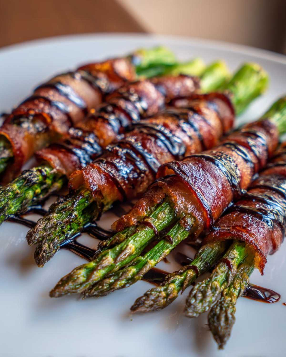 Close-up of cooked Prosciutto Wrapped Asparagus Hearts drizzled with balsamic glaze on a white plate.