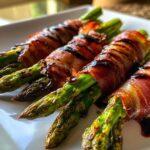 Close-up of perfectly cooked Prosciutto Wrapped Asparagus Hearts drizzled with balsamic glaze on a white plate.