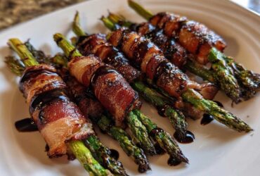 Close-up of cooked Prosciutto Wrapped Asparagus Hearts drizzled with a dark balsamic glaze on a white plate.