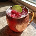A close-up of a frosty copper mug filled with a Raspberry Moscow Mule, garnished with fresh raspberries and a lime wheel.