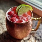Close-up of a vibrant Raspberry Moscow Mule cocktail filled with ice and fresh raspberries, garnished with lime slices in a frosty copper mug.