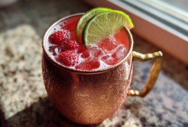 Close-up of a vibrant Raspberry Moscow Mule cocktail filled with ice and fresh raspberries, garnished with lime slices in a frosty copper mug.