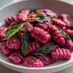 Close-up of vibrant pink Roasted Beet Gnocchi with Sage Butter, garnished with crispy sage leaves and seasoning.